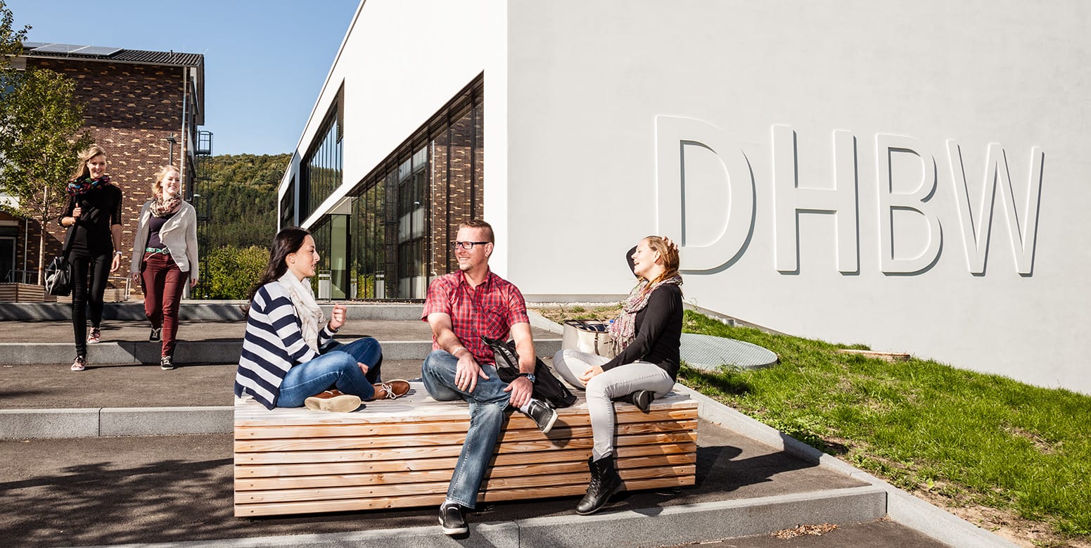 Drei Studierende sitzen vor dem E-Gebäude auf einer Holzbank in der Sonne und unterhalten sich. Im Hintergrund nähern sich zwei Studentinnen. 